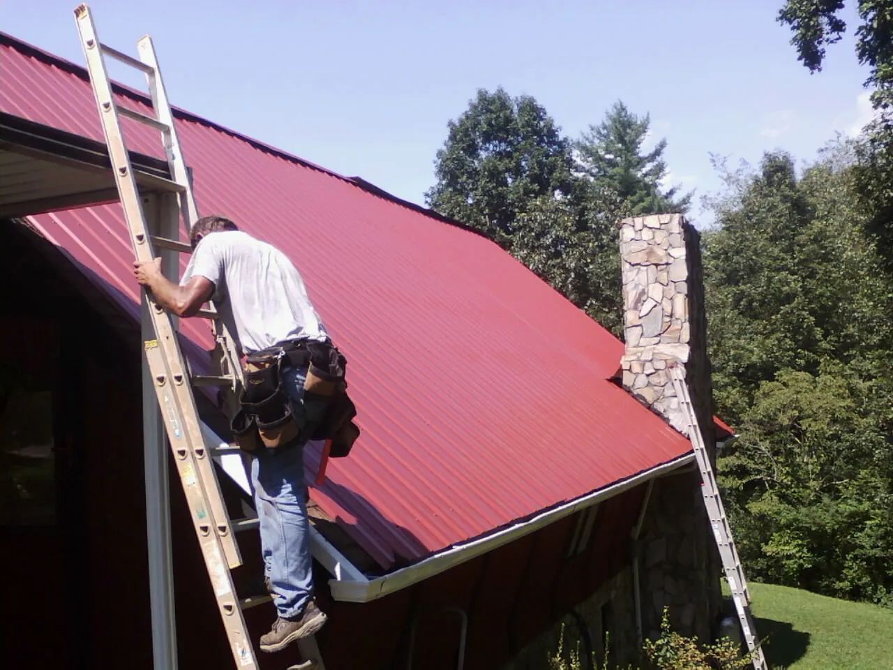Roofer on a ladder beside a red metal roof and stone chimney — Soffit & Fascia Repair by Roofing Brothers in Simsbury